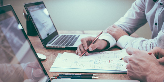 Kooperationen Es lassen sich zwei Personen erkennen, die zusammen vor einem Schreibtisch sitzen und sich austauschen